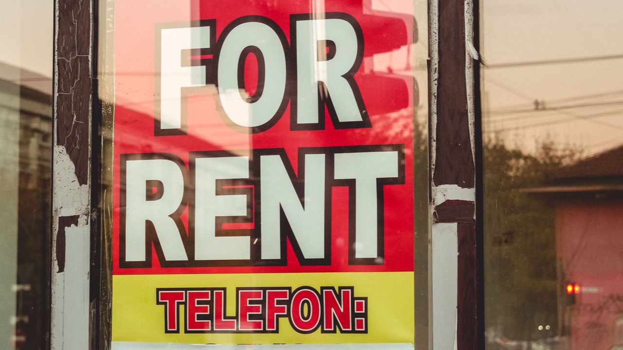 Reflection of a cityscape on a window with a 'For Rent' sign and sunset glow. (Pexels photo)